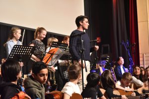 Schülerinnen und Schüler stehen und sitzen auf der Bühne während eines Musikauftritts. Einige halten Notenblätter, während ein Junge im Vordergrund spricht. Im Hintergrund sind weitere Schüler mit Instrumenten zu sehen. Die Bühne ist beleuchtet und es gibt ein großes Projektionsleinwand.