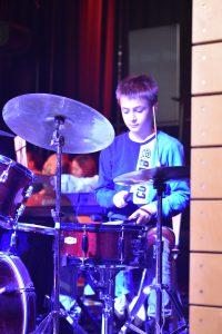 Ein junger Drummer spielt auf einer Bühne. Er sitzt hinter einem Schlagzeug und konzentriert sich auf sein Spiel. Die Bühne ist schwach beleuchtet, mit einem bunten Lichtspiel im Hintergrund, das eine lebendige Atmosphäre schafft.