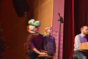 Ein Mann mit einer Yoshi-Mütze sitzt auf einem Stuhl, neben ihm steht ein Junge. Beide schauen in Richtung eines Mikrofons. Im Hintergrund ist ein weiterer Mann zu sehen, der an einem Musikinstrument spielt. Die Szene spielt sich auf einer Bühne mit rotem Vorhang ab.