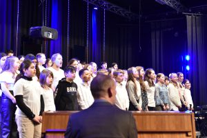 Ein Kinderchor steht auf einer Bühne und singt. Die Sängerinnen und Sänger tragen unterschiedliche Oberteile, überwiegend in hellen Farben. Im Vordergrund sitzt eine Person, die den Chor dirigiert oder begleitet. Die Bühne ist mit blauen Lichtern beleuchtet, und der Hintergrund ist dunkel.