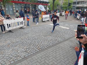 Ein Läufer in sportlicher Kleidung sprintet auf einer gepflasterten Straße, während Zuschauer ihn anfeuern. Im Hintergrund sind bunt gekleidete Personen und Absperrungen zu sehen. Einige Zuschauer halten Smartphones, um den Moment festzuhalten.