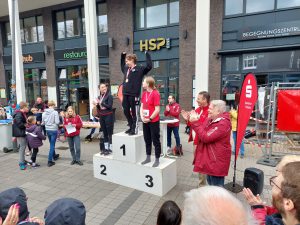 Drei Personen stehen auf einem Podest mit den Stufen 1, 2 und 3. Die Person auf der obersten Stufe hebt einen Pokal in die Luft und wird von Zuschauern applaudiert. Im Hintergrund sind mehrere Personen zu sehen, die ebenfalls applaudieren. Es gibt eine Werbeflagge mit dem Logo einer Bank. Die Szenerie spielt sich draußen vor einem Gebäude ab.