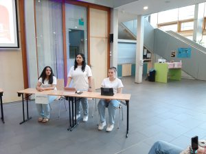 Drei Personen sitzen an einem Tisch in einem hellen Raum. Sie tragen weiße T-Shirts und haben Laptops vor sich. Der Raum hat eine moderne Architektur mit großen Fenstern und einer Treppe im Hintergrund. Eine Person steht aufrecht und spricht, während die anderen beiden aufmerksam zuhören.
