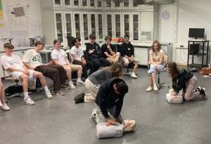 Eine Gruppe von Jugendlichen sitzt in einem Raum, während zwei Personen auf einer Übungspuppe Herz-Lungen-Wiederbelebung (HLW) demonstrieren. Die anderen Teilnehmer beobachten aufmerksam. Der Raum ist hell erleuchtet und hat große Fenster sowie Tische und Stühle.