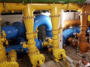 Farbige Rohrleitungen in einer industriellen Umgebung, darunter gelbe, blaue und orange Rohre, die miteinander verbunden sind. Im Vordergrund ist ein Teil eines menschlichen Körpers sichtbar, der an der Arbeit ist.