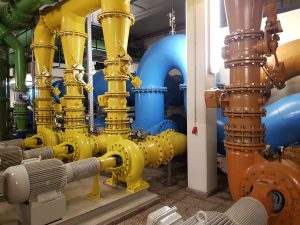 Blick auf eine industrielle Rohrleitungsanlage mit großen, farbigen Rohren in Gelb, Blau und Orange. Im Vordergrund sind mehrere Motoren sichtbar, die zur Steuerung der Rohrsysteme dienen. Die Umgebung wirkt sauber und gut beleuchtet.