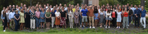 Gruppenfoto von etwa 100 Personen im Freien, stehend vor einem grünen Hintergrund. Die Personen tragen verschiedene Kleidungsstile, von formell bis lässig, und lächeln in die Kamera. Es sind sowohl Männer als auch Frauen in unterschiedlichen Altersgruppen zu sehen.