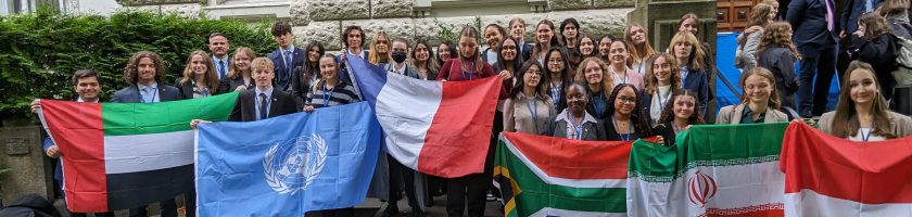 Eine Gruppe von etwa 50 Personen steht vor einem Gebäude und hält verschiedene Flaggen in den Händen, darunter die Flaggen von Frankreich, den Vereinten Nationen, Südafrika, Iran und anderen. Die Teilnehmerinnen und Teilnehmer lächeln und posieren für das Foto. Das Wetter scheint schön zu sein, und die Umgebung ist grün.