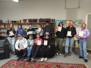 Gruppe von zwölf Jugendlichen, die in einem Raum stehen und sitzen, während sie Bücher in den Händen halten. Im Hintergrund sind Regale mit weiteren Büchern zu sehen. Der Raum hat einen Teppichboden und eine gemütliche Atmosphäre.
