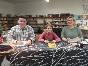 Drei Personen sitzen an einem Tisch in einer Bibliothek. Links ein junger Mann in einem karierten Hemd, in der Mitte ein Junge in einem roten Hoodie und rechts eine Frau in einem grünen Oberteil. Auf dem Tisch liegt ein Buch und es sind Lichterketten dekoriert. Im Hintergrund sind Regale mit Büchern sichtbar.