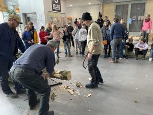 Eine Gruppe von Menschen beobachtet eine Holzverarbeitungsdemonstration in einem Innenraum. Zwei Personen sind aktiv am Arbeiten, während die Zuschauer in einem Halbkreis darum stehen. Einige der Zuschauer sitzen auf dem Boden, andere stehen. Der Boden ist mit Holzspänen bedeckt, und die Atmosphäre wirkt informell und lebhaft.