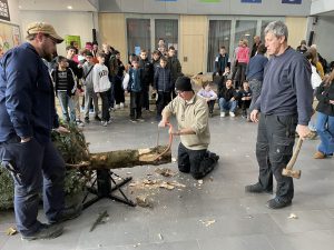 Drei Männer arbeiten in einem Innenraum, um einen Baumstamm zu bearbeiten. Einer von ihnen sitzt auf dem Boden und schnitzt mit einem Werkzeug, während die anderen beiden stehen und zusehen. Im Hintergrund sind viele Menschen, darunter Kinder und Jugendliche, die interessiert zuschauen. Der Boden ist mit Spänen und Holzstücken bedeckt.