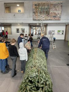 Eine Gruppe von Menschen steht in einem Innenraum um einen großen, liegenden Weihnachtsbaum herum. Im Hintergrund sind Leitern und ein Wandgemälde zu sehen. Einige Personen arbeiten an dem Baum, während andere zuschauen. Der Raum wirkt hell und freundlich.