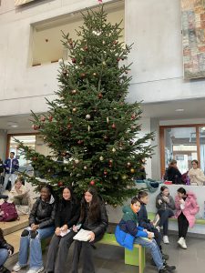 Eine große, geschmückte Tanne steht in einem hellen Raum. Vor dem Baum sitzen mehrere Kinder und Jugendliche auf einer Bank. Einige von ihnen lächeln und unterhalten sich, während im Hintergrund weitere Personen sichtbar sind, die sich im Raum aufhalten. Der Raum wirkt festlich und einladend.