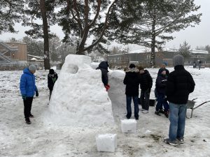 Eine Gruppe von sieben Kindern steht auf einem verschneiten Platz und baut gemeinsam einen Schneemann oder eine Schneehütte. Einige Kinder tragen Winterjacken und Mützen, während sie mit Schnee arbeiten. Im Hintergrund sind Bäume und eine Schulgebäude zu sehen. Es ist ein bewölkter Tag mit leichtem Schneefall.