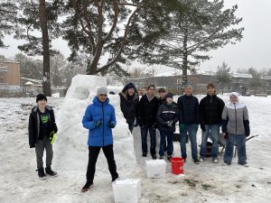 Gruppe von sieben Personen steht im Schnee, umgeben von Bäumen. Im Hintergrund ist eine große Schneeskulptur zu sehen. Die Personen tragen winterliche Kleidung, einige halten Eimer oder andere Utensilien in der Hand. Es schneit leicht.