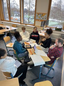Sechs Personen sitzen in einem Klassenraum an einem Tisch, umgeben von Fenstern. Sie diskutieren und arbeiten an Laptops und Notizen. Die Gruppe besteht aus drei Frauen und drei Männern, die verschiedene Kleidung tragen. Der Raum wirkt hell und einladend.