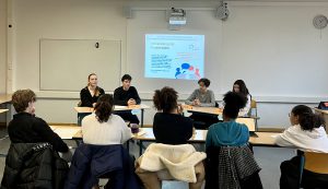 Eine Gruppe von Personen sitzt an einem Tisch in einem Klassenraum. Im Vordergrund sind mehrere Personen sichtbar, die auf eine Präsentation an der Wand schauen. Auf der Präsentation sind Grafiken und Text zu sehen, die möglicherweise ein Thema oder eine Diskussion darstellen. Der Raum ist hell erleuchtet und hat eine einfache Ausstattung.