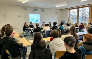Eine Gruppe von Schülern sitzt in einem Klassenraum, während eine Diskussion oder Präsentation stattfindet. Im Vordergrund sind mehrere Schüler zu sehen, die aufmerksam auf die Referenten an einem Tisch achten. An der Wand ist ein Projektor mit einer Präsentation zu sehen. Der Raum hat große Fenster, die Tageslicht hereinlassen.