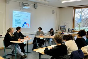 In einem Klassenzimmer sitzen fünf Personen an Tischen. An der Wand ist ein Projektor mit einer Präsentation zu sehen. Die Anwesenden sind in ein Gespräch vertieft, während einige Notizen machen. Es gibt Fenster, durch die Tageslicht hereinfällt.