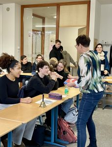 Eine Lehrerin steht vor einer Gruppe von Schülerinnen und Schülern in einem Klassenzimmer. Einige Schüler sitzen an Tischen und schauen aufmerksam zur Lehrerin, während andere im Hintergrund stehen. Auf einem Tisch steht eine kleine Glocke und eine Flasche Wasser. Die Atmosphäre wirkt freundlich und interessiert.