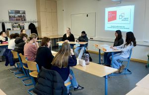Eine Gruppe von Schülerinnen und Schülern sitzt in einem Klassenzimmer an Tischen. Vor ihnen stehen drei Personen, die eine Präsentation halten. Im Hintergrund ist eine Leinwand mit einem Präsentationsinhalt sichtbar. Die Wände sind hell gestrichen, und es gibt einige Schränke sowie ein Plakat an der Wand. Die Atmosphäre ist konzentriert und lehrreich.