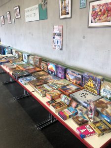 Ein Tisch mit einer Vielzahl von Brettspielen, die ordentlich aufgereiht sind. Im Hintergrund sind Bilder an der Wand zu sehen. Die Tischoberfläche ist hell, und der Boden ist dunkel. Es gibt viel Tageslicht in dem Raum.
