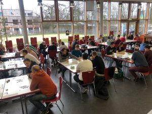 Eine Gruppe von Menschen sitzt an Tischen in einem großen Raum. Die Teilnehmer sind in konzentrierter Unterhaltung oder beim Spielen von Brettspielen. Der Raum hat große Fenster, die viel Tageslicht hereinlassen, und im Hintergrund sind weitere Tische und Stühle zu sehen.