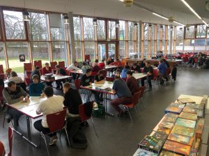 Eine Gruppen von Menschen sitzt an Tischen in einem hellen Raum mit großen Fenstern, die auf eine grüne Außenansicht blicken. Auf den Tischen liegen verschiedene Spiele. Die Atmosphäre wirkt gesellig und einladend, während die Teilnehmer sich unterhalten und miteinander interagieren.