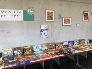 Ein Tisch mit einer Vielzahl von Brettspielen ist in einem Raum aufgestellt. Im Hintergrund sind an einer Betonwand verschiedene Bilder und Schilder angebracht, darunter ein Schild mit der Aufschrift 