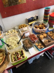 Ein Tisch mit verschiedenen Speisen und Snacks. Darunter sind Salate, Wurstwaren, Gebäck, Früchte und Desserts in verschiedenen Schalen und Platten angerichtet. Einige Schalen sind mit Beschriftungen versehen. Im Hintergrund sind bunte Becher zu sehen.