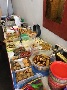 Ein langer Tisch ist mit verschiedenen Speisen und Snacks gedeckt. Es sind mehrere Schalen und Behälter mit salzigen und süßen Leckereien, darunter Kekse, Gebäck, kleine Häppchen und Obst. Bunte Teller und Besteck sind ebenfalls auf dem Tisch platziert. Der Hintergrund zeigt eine einfache Wand und einen Spiegel.