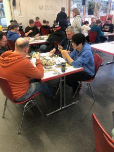 Eine Gruppe von Personen sitzt an Tischen in einem Raum, der für soziale Aktivitäten genutzt wird. Einige Teilnehmer sind beschäftigt, Karten oder Spielmaterialien zu betrachten, während andere miteinander sprechen. Die Umgebung ist hell und freundlich, mit Stühlen in verschiedenen Farben.