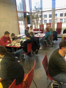 Eine Gruppe von Menschen sitzt an Tischen in einem hellen Raum mit großen Fenstern. Einige Personen lesen oder arbeiten an Projekten, während andere miteinander sprechen. Die Tische sind mit verschiedenen Materialien und Gegenständen bedeckt. Im Hintergrund sind rote Stühle gestapelt zu sehen.