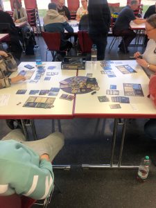 Zwei Personen sitzen an einem Tisch, der mit verschiedenen Spielkarten und einem Spielbrett bedeckt ist. Im Hintergrund sind weitere Personen zu sehen, die ebenfalls an Tischen sitzen. Die Umgebung wirkt freundlich und gesellig, und es sind Getränke auf dem Tisch vorhanden.
