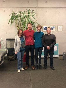 Vier Personen stehen lächelnd vor einer Wand mit Pflanzen und einem Tisch. Die erste Person links trägt einen grauen Cardigan, die zweite einen roten Pullover, die dritte einen blauen Pullover und die vierte eine graue Sweatshirt. Im Hintergrund sind einige Schilder und ein Tisch zu sehen.