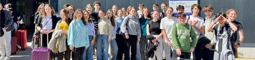 Gruppe von etwa 30 Personen, die vor einem Gebäude stehen, einige mit Gepäck. Sie lächeln und posieren für ein Foto, während die Sonne scheint.