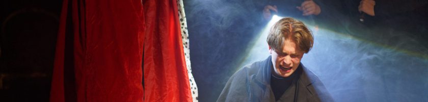 Ein Mann steht auf einer Bühne, umgeben von einem Nebel und beleuchtet von einem Scheinwerfer. Im Vordergrund ist ein roter Vorhang zu sehen. Der Mann zeigt eine ausdrucksvolle Miene, die möglicherweise Emotionen wie Freude oder Erstaunen widerspiegelt.