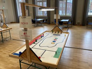 Ein Tisch in einem großen Raum mit Holzfußboden. Auf dem Tisch befindet sich ein Spielfeld mit einer gezeichneten Strecke, die von verschiedenen bunten Figuren (Rot, Blau, Gelb, Grün) umrandet ist. Eine Beschilderung mit der Aufschrift 