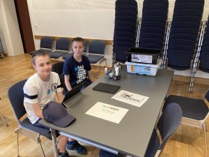 Zwei Jungen sitzen an einem Tisch in einem Raum mit gestapelten Stühlen im Hintergrund. Auf dem Tisch befinden sich zwei Tablets, einige Dokumente und ein kleiner Behälter. Die Jungen schauen in die Kamera und scheinen an einem Projekt oder einer Aktivität beteiligt zu sein.