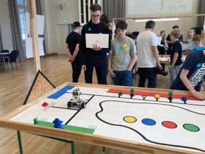 Zwei Jugendliche stehen an einem Tisch, auf dem ein Roboter und eine farbige Strecke platziert sind. Der ältere Junge hält ein Tablet in der Hand, während der jüngere Junge auf den Tisch schaut. Im Hintergrund sind weitere Personen und ein Bildschirm sichtbar.