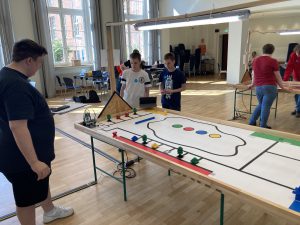 Drei Personen arbeiten an einem großen, rechteckigen Tisch, der mit einer farbigen, gezeichneten Spielfläche ausgestattet ist. Auf dem Tisch befinden sich verschiedene Objekte und Roboter, die für ein Projekt oder einen Wettbewerb verwendet werden. Im Hintergrund sind weitere Personen und Tische zu sehen, die auf eine aktive Lernumgebung hinweisen. Das Licht fällt durch große Fenster und schafft eine helle Atmosphäre im Raum.