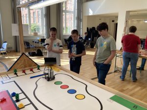 Drei Jugendliche stehen um einen großen Tisch mit einem Spielbrett, das eine farbige Strecke und verschiedene Spielsteine zeigt. Sie betrachten einen Laptop oder ein Tablet, während ein weiterer Jugendlicher im Hintergrund an einem anderen Tisch arbeitet. Der Raum hat große Fenster mit Tageslicht und eine helle Holzfußboden.