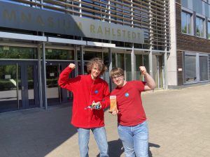Zwei Jugendliche stehen vor dem Gebäude des Gymnasiums Rahlstedt. Der Junge links trägt einen roten Pullover und zeigt mit einer Faust nach oben, während er einen kleinen Gegenstand in der anderen Hand hält. Das Mädchen rechts trägt ein rotes T-Shirt und hält eine Auszeichnung oder einen Preis in der Hand. Beide lächeln und wirken erfreut. Im Hintergrund ist der Eingang des Gymnasiums mit dem Schriftzug „Gymnasium Rahlstedt“ zu sehen.