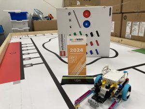 Ein LEGO-Roboter steht auf einer Spielfläche mit einer markierten Strecke. Im Hintergrund ist eine große Box mit bunten Symbolen zu sehen, und ein Schild mit der Aufschrift 