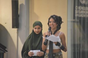 Zwei Frauen stehen nebeneinander auf einer Bühne. Die Frau links trägt ein grünes Kopftuch und ein einfaches Outfit, während die Frau rechts ein aufregenderes Outfit mit einem Oberteil ohne Ärmel und einer Krawatte trägt. Sie hält ein Mikrofon und liest von einem Blatt Papier ab. Im Hintergrund sind unscharfe Elemente einer Stadtumgebung zu sehen.