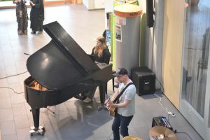 Zwei Musiker treten in einem Raum auf. Eine Person spielt am Klavier, während die andere Gitarre spielt. Im Hintergrund sind weitere Menschen zu sehen, die zuschauen. Der Raum hat helle Fliesen und eine offene Atmosphäre.
