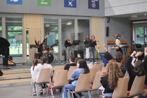 Eine Band spielt auf einer Bühne in einem Innenraum, während eine Gruppe von Menschen im Publikum zuschaut und begeistert applaudiert. Im Hintergrund sind Musikinstrumente wie ein Schlagzeug und Tasteninstrumente sichtbar. Die Wände sind mit verschiedenen Fahnen oder Symbolen dekoriert.