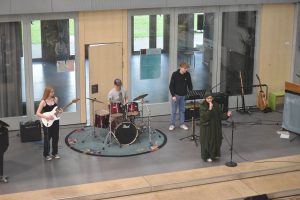Eine Band spielt auf einer Bühne in einem modernen Raum. Eine Sängerin mit einem langen, grünen Kleid steht am Mikrofon, während ein Gitarrist, ein Schlagzeuger und ein weiterer Musiker im Hintergrund spielen. Es gibt verschiedene Musikinstrumente und die Wände sind mit Fenstern versehen, durch die Tageslicht strömt.