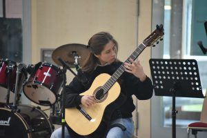 Junge Frau spielt Gitarre auf einer Bühne, im Hintergrund sind Schlaginstrumente zu sehen. Sie sitzt auf einem Stuhl und konzentriert sich auf ihr Spiel. Ein Notenständer steht neben ihr.